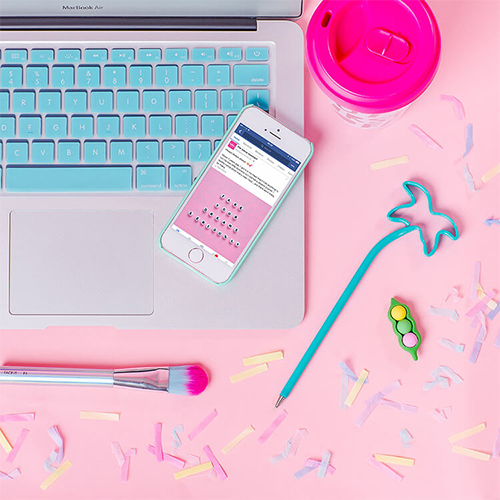 Bureau rose pastel saupoudré de confettis sur lequel est posé un MacBook au clavier bleu, un Iphone, un stylo bleu en forme de palmier, un mug rose fushia et un pinceau de maquillage rose